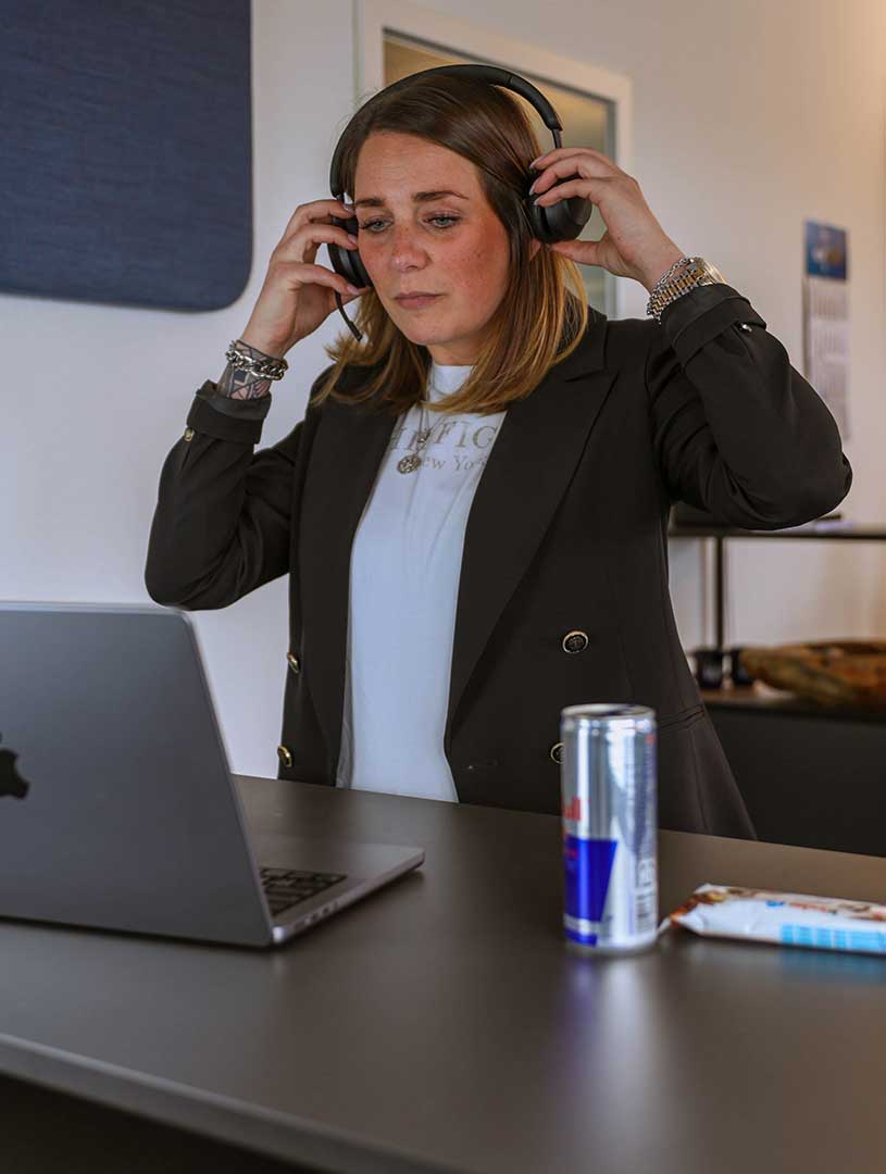 Eine Kollegin trägt Kopfhörer und arbeitet konzentriert an ihrem Laptop. Auf dem Tisch stehen ein Energydrink und ein Snack.
